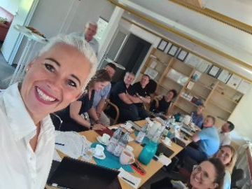 Lächende Personen sitzen an einem langen Holztisch mit Kaffeetassen und Laptops. Eine Frau steht im Vordergrund lächend und macht ein Selfie der Gruppe.