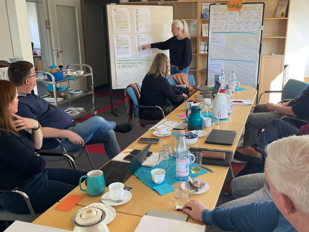 Foto einer Besprechung in einem Konferenzraum mit mehreren Personen, die einem Vortrag an einem Flipchart folgen. Auf dem Tisch sind Laptops, Notizbücher und Getränke zu sehen. Die Szene wirkt professionell und fokussiert.